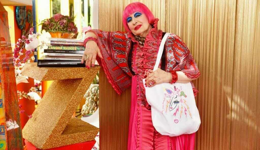 Zandra Rhodes with electrical tote bag wearing all pink and leaning on a book shelf