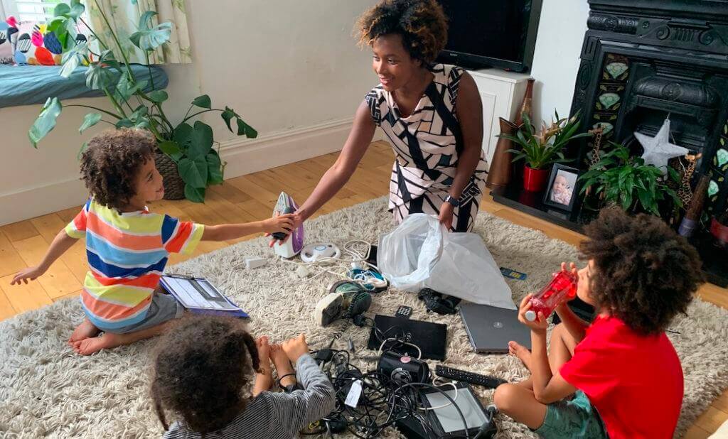 A family (one dark skinned woman and three medium dark skinned children) sitting on a rug in a living room sorting electricals for recycling.