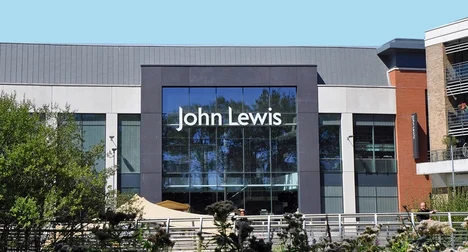 Entrance of a John Lewis store with shiny glass windows reflecting the blue sky and trees surrounding the building.