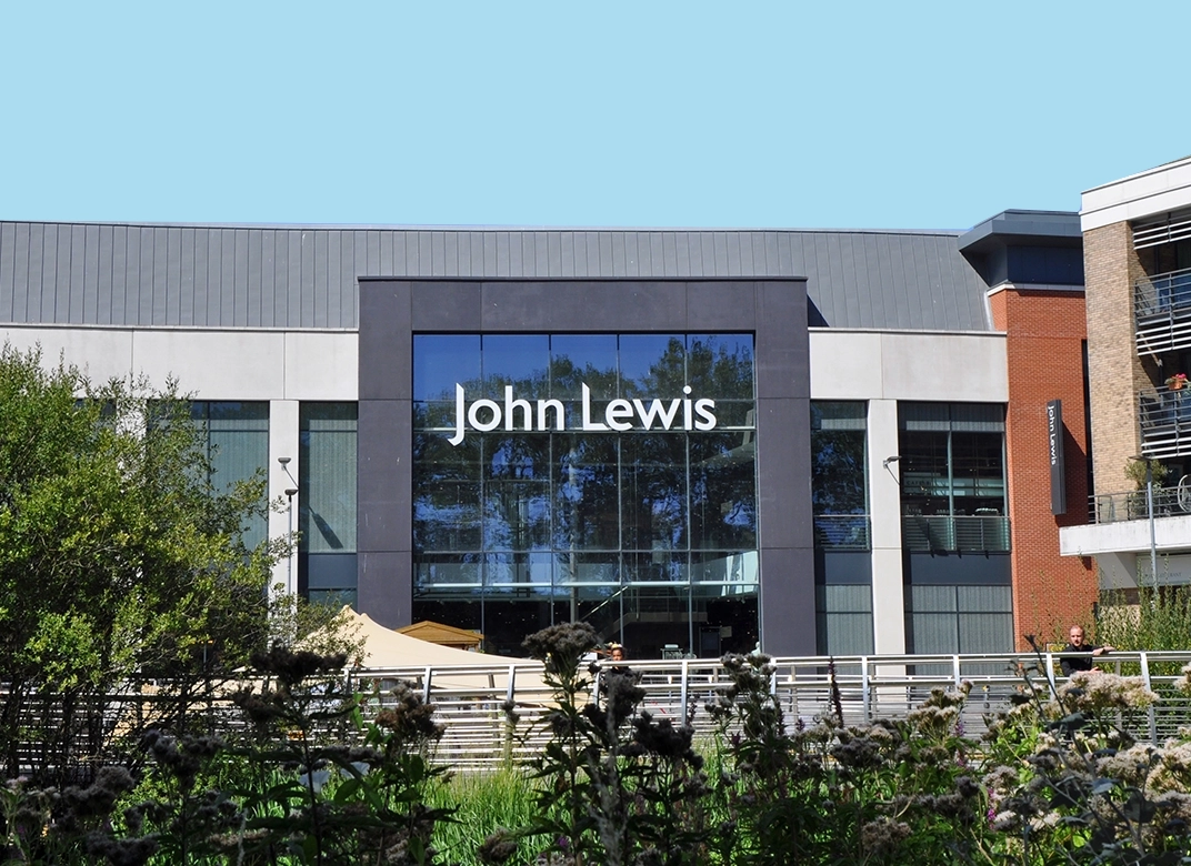 Entrance of a John Lewis store with shiny glass windows reflecting the blue sky and trees surrounding the building.