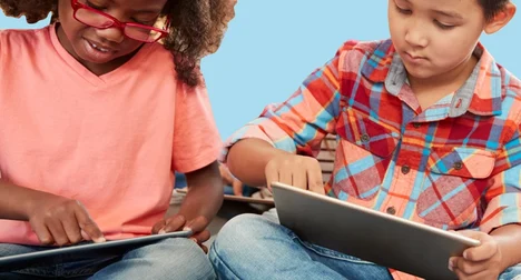 Two children sitting cross legged using tablets against a bright blue background.