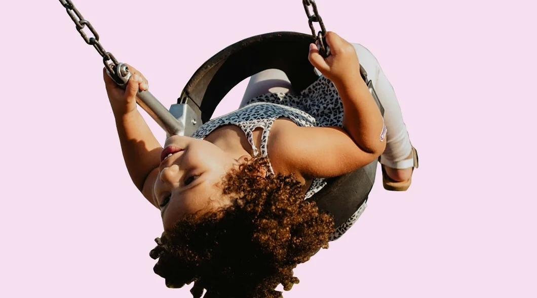 A medium skinned child on a tyre swing, mid-swing upside down, against a pink background.