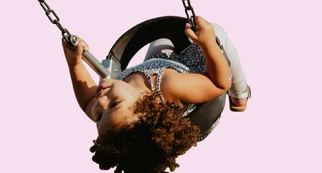 A medium skinned girl on a swing, mid-swing upside down, against a pink background.