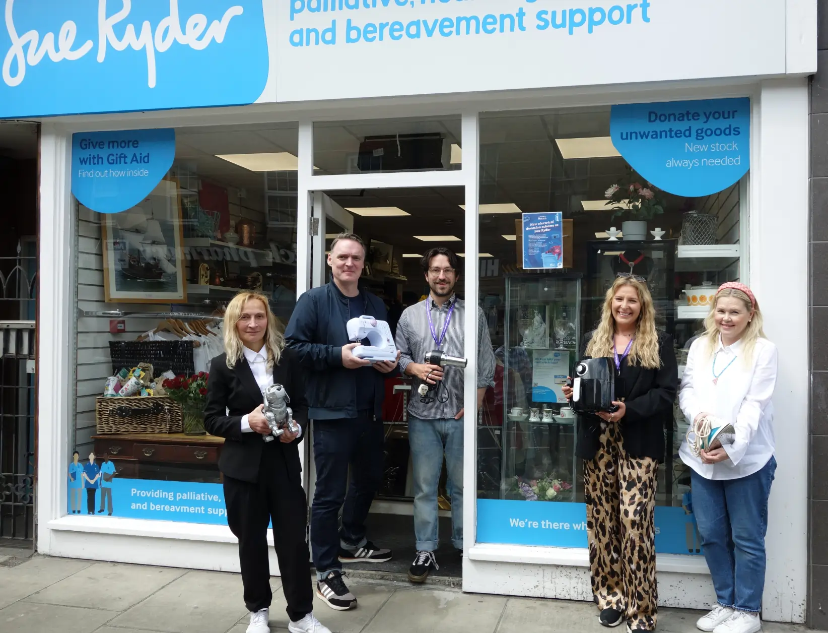 three pale skinned women and two pale skinned men standing in the doorway of a Sue Ryder charity shop