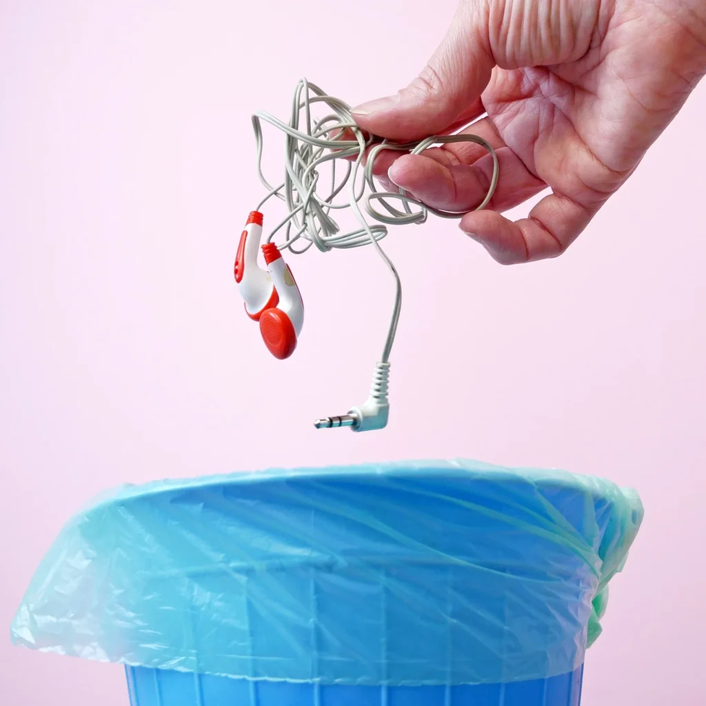 a pale skinned hand throwing someearbuds into a blue waste bin on a plain pink background